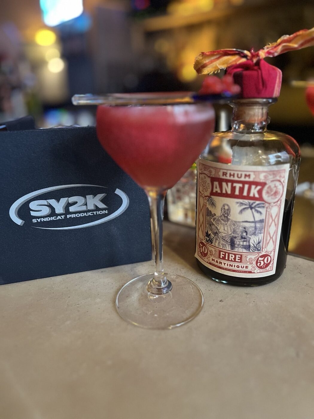 The Ultimate Sour servi au Syndicat, à côté d'une bouteille d'ANTIK Fire, sur un tapis de bar SY2K